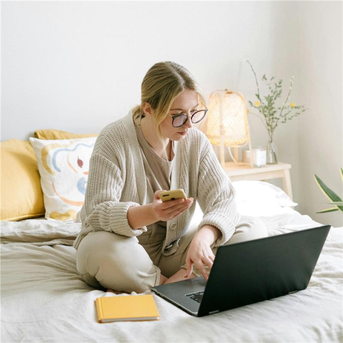 woman sits on her bed with a laptop in front of her and her phone in her right hand. She is reviewing her tax information on her laptop and phone simultaneously.