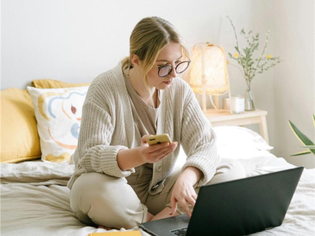 woman sits on her bed with a laptop in front of her and her phone in her right hand. She is reviewing her tax information on her laptop and phone simultaneously.