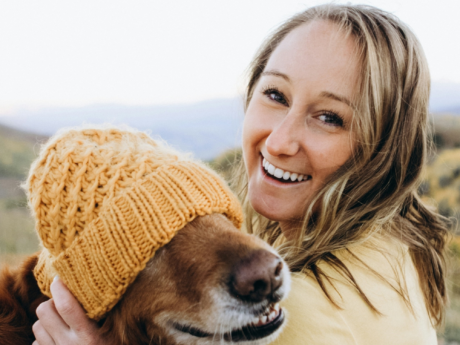 Smiling woman sitting outdoors holding a dog wearing a yellow hat