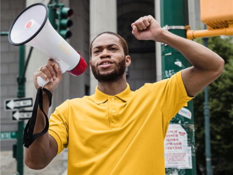 Man with a beard and wearing a yellow shirt holds a megaphone and raises one arm in the air.