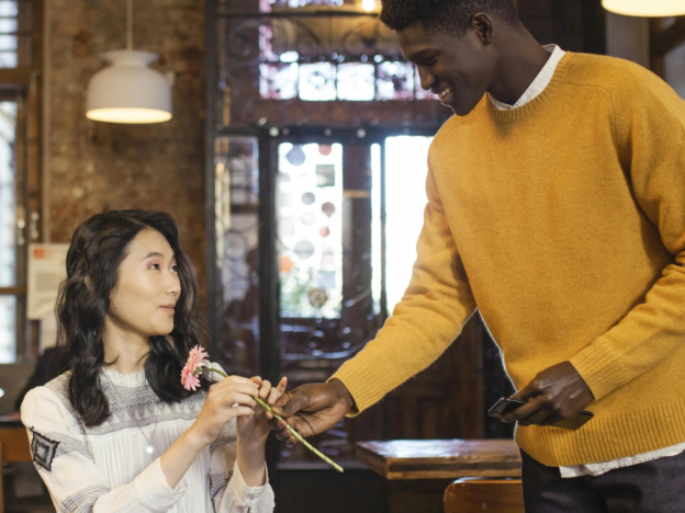 An image of a man and woman at a cafe. The woman is sitting at a table with a cup of coffee, and the man has approached her and is standing next to her giving her a flower.