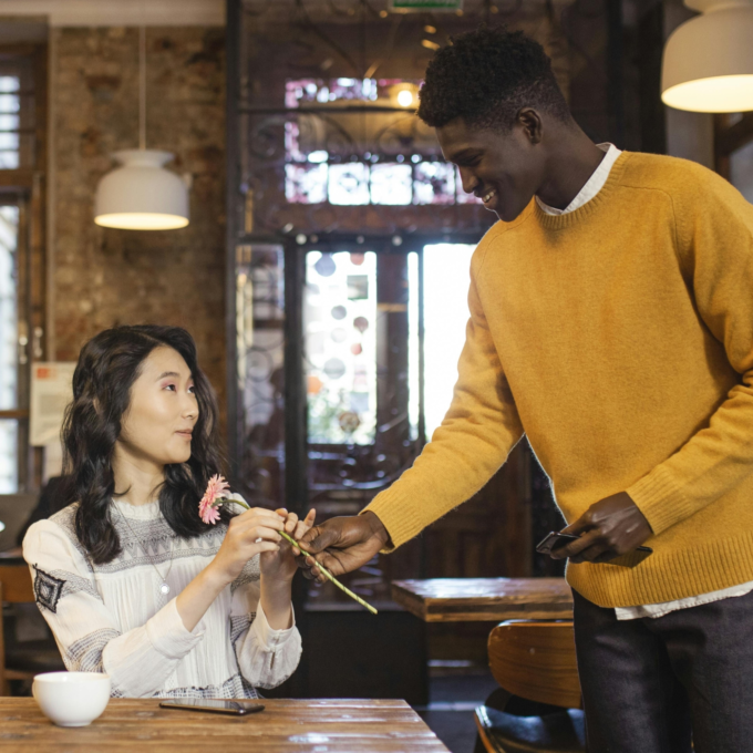 An image of a man and woman at a cafe. The woman is sitting at a table with a cup of coffee, and the man has approached her and is standing next to her giving her a flower.