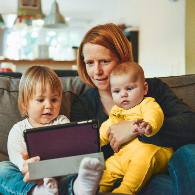 Middle-aged female sitting on a couch holding an infant child, while a toddler sits next to her holding a tablet.