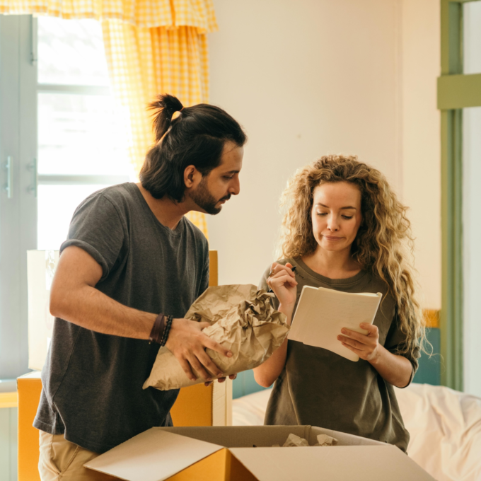 A man and a woman are unpacking boxes in a room with large windows and yellow curtains. The man is removing a wrapped item from the box, and the woman is looking at a notebook that she is holding. They are surrounded by cardboard boxes.