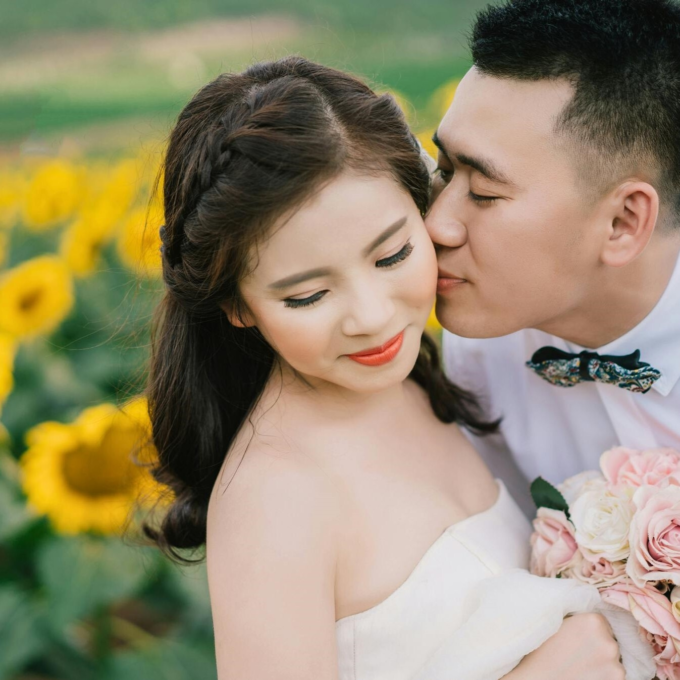 A couple embracing for a wedding photo in a sunflower field. The bride is in a white dress holding a bouquet, the groom is in a white shirt and bowtie.