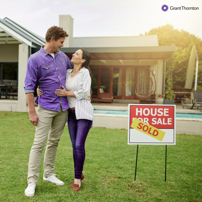 Couple standing on a lawn in front of a modern house with a “House for Sale” sign marked “Sold.”