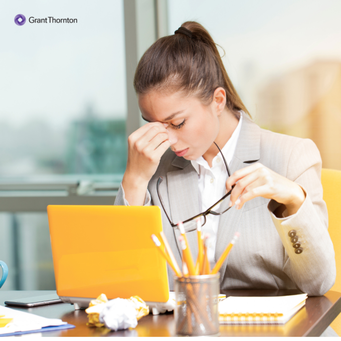 Person looking stressed sitting at a desk with a laptop, holding eyeglasses in one hand and touching their forehead while working in a bright office setting.