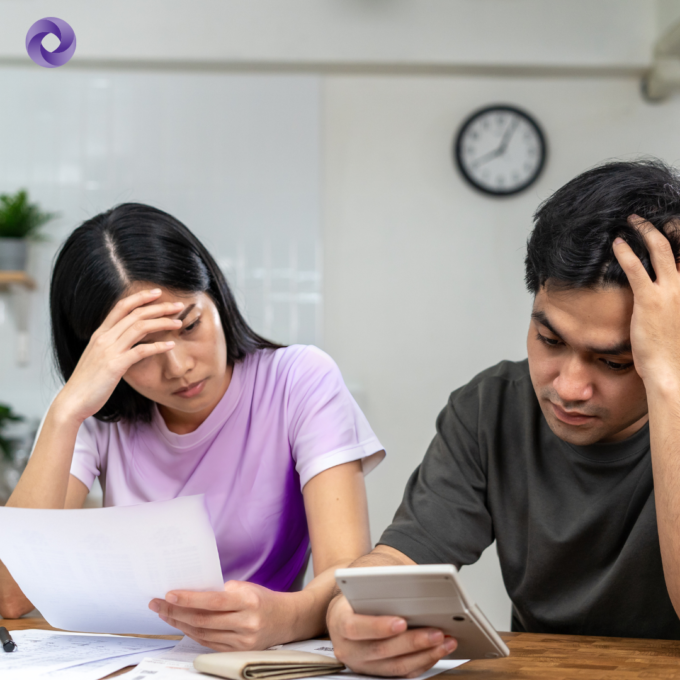 A couple sits at a table reviewing bills and using a calculator, appearing stressed while looking over financial documents.