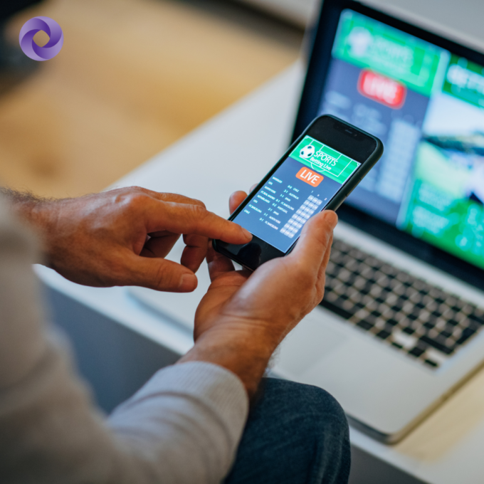 Hands holding a smartphone displaying a live sports betting app, with additional betting information shown on a laptop screen in the background.