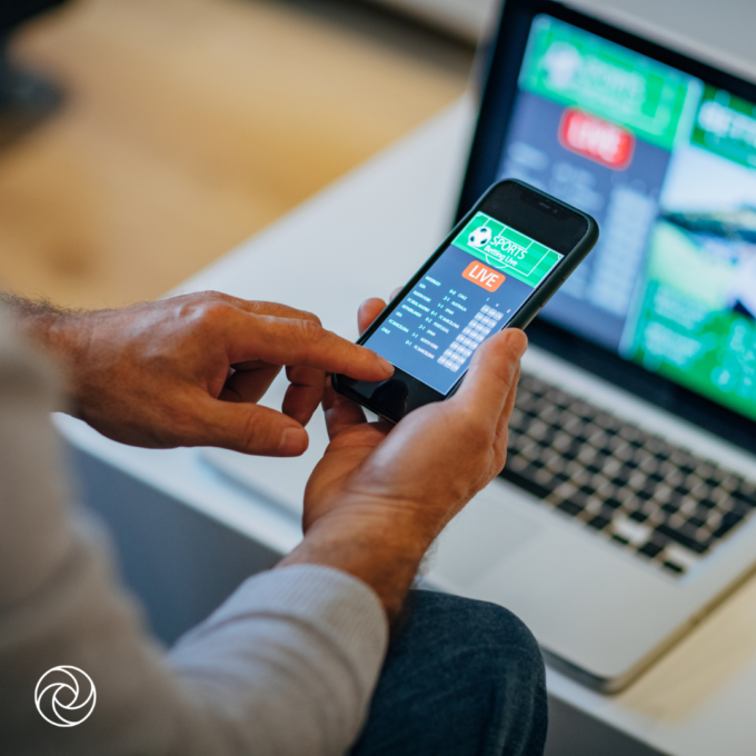 Hands holding a smartphone displaying a live sports betting app, with additional betting information shown on a laptop screen in the background.