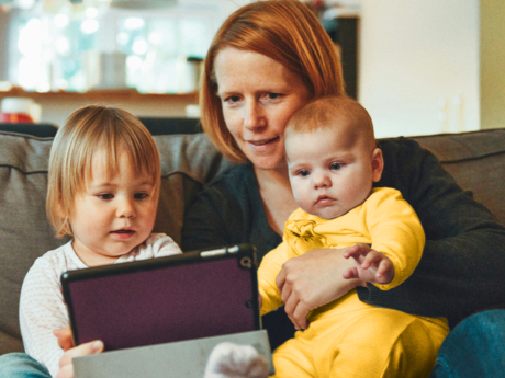 Middle-aged female sitting on a couch holding an infant child, while a toddler sits next to her holding a tablet.