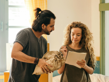A man and a woman are unpacking boxes in a room with large windows and yellow curtains. The man is removing a wrapped item from the box, and the woman is looking at a notebook that she is holding. They are surrounded by cardboard boxes.