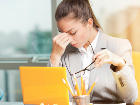 Person looking stressed sitting at a desk with a laptop, holding eyeglasses in one hand and touching their forehead while working in a bright office setting.