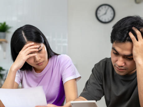 A couple sits at a table reviewing bills and using a calculator, appearing stressed while looking over financial documents.