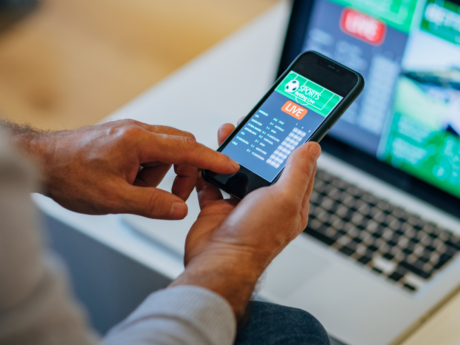 Hands holding a smartphone displaying a live sports betting app, with additional betting information shown on a laptop screen in the background.