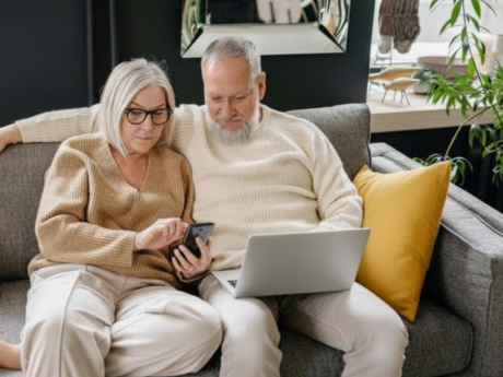 An older aged couple sitting together on the couch looking at their laptop and phone