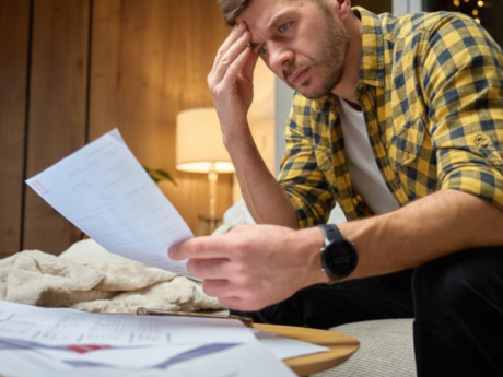 Person looking down at bills with head in hand, surrounded by paperwork and an open laptop.
